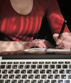 A person wearing a red shirt is writing in a notebook with a pen. In the foreground, the keyboard of a laptop is visible, suggesting a workspace setting. Soft light and shadows create a relaxed atmosphere.