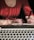 A person wearing a red shirt is writing in a notebook with a pen. In the foreground, the keyboard of a laptop is visible, suggesting a workspace setting. Soft light and shadows create a relaxed atmosphere.
