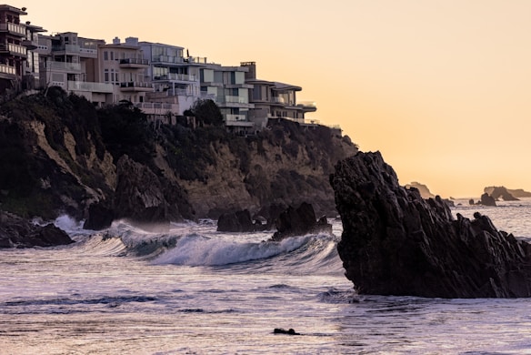 A coastal scene with modern, multi-story houses perched atop a rugged cliff, overlooking a rocky shoreline and crashing waves. The buildings display large windows and balconies, suggesting luxury and a scenic view. The sky has a warm, golden hue, indicative of either sunrise or sunset, casting a soft light over the water.