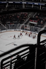 A hockey game taking place in an indoor arena with a mostly empty audience. Players in red and white uniforms are actively participating on the ice rink. The area is surrounded by transparent protective barriers and advertisements are visible along the boards.