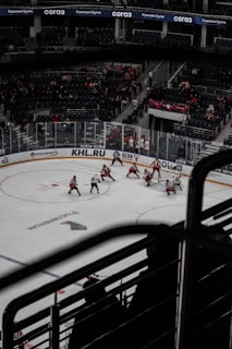 A hockey game taking place in an indoor arena with a mostly empty audience. Players in red and white uniforms are actively participating on the ice rink. The area is surrounded by transparent protective barriers and advertisements are visible along the boards.