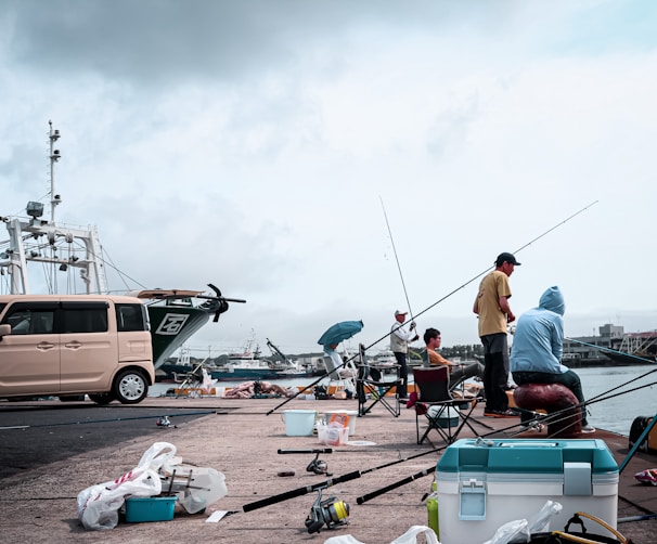 Several people are fishing on a pier by the water. They are surrounded by various fishing gear, chairs, and coolers. A small beige van is parked nearby, and fishing boats are visible in the background. The sky is overcast, suggesting a cloudy day.