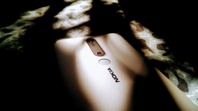 A Nokia smartphone laying on a surface with a pattern. The phone is partially illuminated by light, highlighting the brand and camera area. The surrounding area is dark with blurred and out-of-focus patterned fabric.