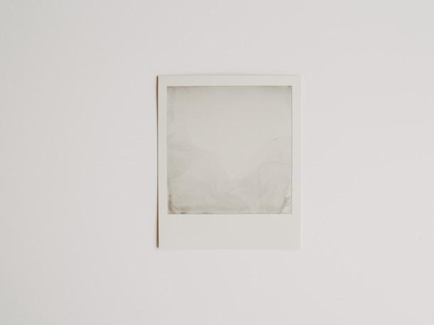 A blank Polaroid photograph is positioned against a plain white background. The image inside the photograph is not developed or visible, resulting in an empty, grey-ish square within the white frame.