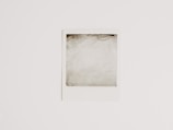 A blank Polaroid-style photograph with a white border lies flat against a light, neutral background. The photo area is empty, showing only a washed-out pale gray color.