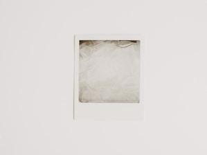 A blank Polaroid-style photograph with a white border lies flat against a light, neutral background. The photo area is empty, showing only a washed-out pale gray color.