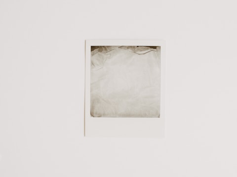 A blank Polaroid-style photograph with a white border lies flat against a light, neutral background. The photo area is empty, showing only a washed-out pale gray color.