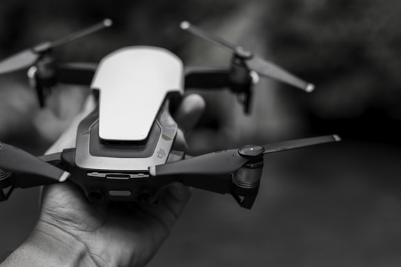 A close-up of a small quadcopter drone held in a person's hand, featuring sleek design and prominent blades. The background is blurred, emphasizing the focus on the drone.