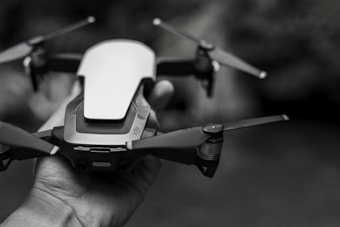 A close-up of a small quadcopter drone held in a person's hand, featuring sleek design and prominent blades. The background is blurred, emphasizing the focus on the drone.