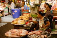 A bustling market scene with women selling various fresh goods. Large fish are displayed in the foreground, while a variety of poultry can be seen on a table. The women are engaged in conversation, reflecting a lively and communal environment. Various fresh produce and containers are visible in the background, creating a vibrant marketplace atmosphere.