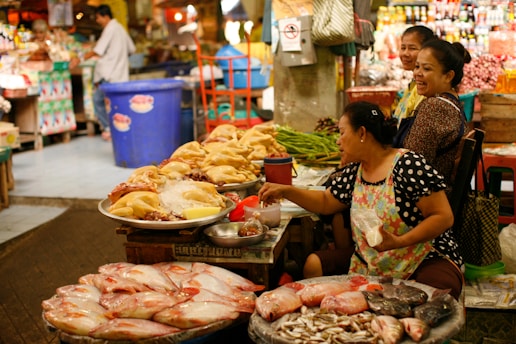 A bustling market scene with women selling various fresh goods. Large fish are displayed in the foreground, while a variety of poultry can be seen on a table. The women are engaged in conversation, reflecting a lively and communal environment. Various fresh produce and containers are visible in the background, creating a vibrant marketplace atmosphere.