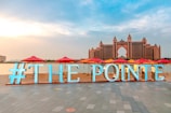 A large, illuminated sign reads '#THE POINTE' on a promenade by the water. In the distance, a grand hotel with distinctive architecture looms, featuring a central archway and multiple turrets. The scene is adorned with red and yellow umbrellas lining the waterfront.