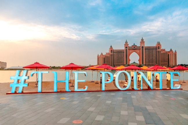 A large, illuminated sign reads '#THE POINTE' on a promenade by the water. In the distance, a grand hotel with distinctive architecture looms, featuring a central archway and multiple turrets. The scene is adorned with red and yellow umbrellas lining the waterfront.