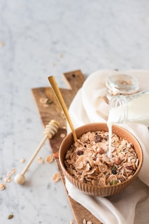 A wooden bowl filled with various flakes and grains is placed on a wooden board with milk being poured from a glass bottle. A golden spoon rests inside the bowl, and a honey dipper lies nearby. The scene is set on a marble surface with a white cloth partially covering the board.