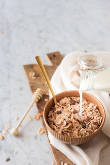A wooden bowl filled with various flakes and grains is placed on a wooden board with milk being poured from a glass bottle. A golden spoon rests inside the bowl, and a honey dipper lies nearby. The scene is set on a marble surface with a white cloth partially covering the board.