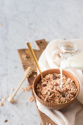 A wooden bowl filled with various flakes and grains is placed on a wooden board with milk being poured from a glass bottle. A golden spoon rests inside the bowl, and a honey dipper lies nearby. The scene is set on a marble surface with a white cloth partially covering the board.