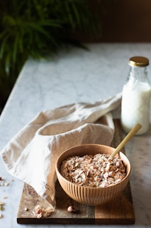 A wooden bowl filled with a mixture of granola and nuts is placed on a wooden cutting board. Next to it is a light-colored cloth draped casually, enhancing the rustic aesthetic. A closed glass bottle filled with milk stands nearby on the marble surface, completing the breakfast setting. In the background, blurred greenery adds to the natural and serene ambiance.