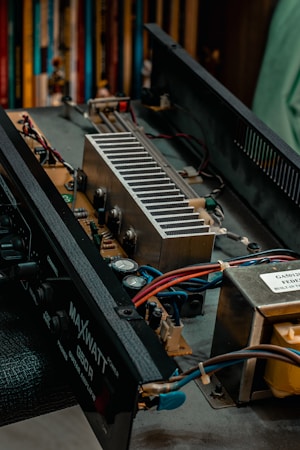 An exposed electronic device with visible internal components including capacitors, heat sinks, and wires. The focus is on the internal layout of what appears to be a guitar amplifier. Several colored wires are connected to different parts of the circuit, and there are printed labels on some components.
