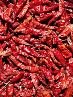 A close-up of numerous dried red chili peppers creates a textured and vibrant display. The chilies are densely packed, showcasing their wrinkled surfaces and deep red hues with occasional darker and lighter highlights.