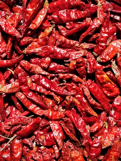 A close-up of numerous dried red chili peppers creates a textured and vibrant display. The chilies are densely packed, showcasing their wrinkled surfaces and deep red hues with occasional darker and lighter highlights.