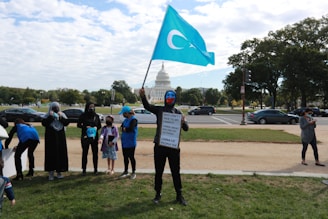 A group of people is gathered on a lawn with the United States Capitol building in the background. One person is holding a blue flag with a white crescent and star. Another person is holding a sign with a message about family and China. The scene appears to be a protest or demonstration.