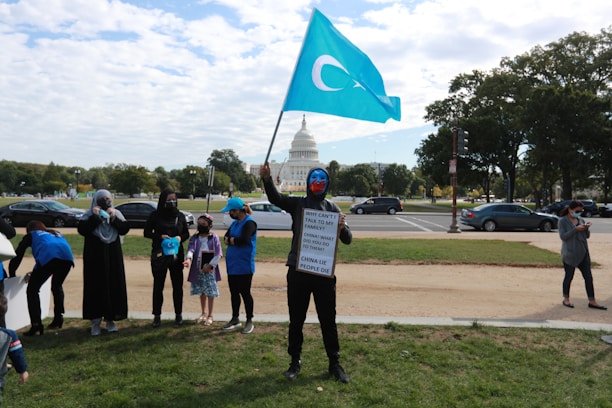 A group of people is gathered on a lawn with the United States Capitol building in the background. One person is holding a blue flag with a white crescent and star. Another person is holding a sign with a message about family and China. The scene appears to be a protest or demonstration.