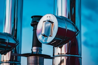 A close-up view of metallic industrial pipes reflecting light against a bright blue sky. The shiny surfaces highlight the cylindrical shapes and the detailed structure of the pipes. One prominent pipe with a circular cap is central to the image.