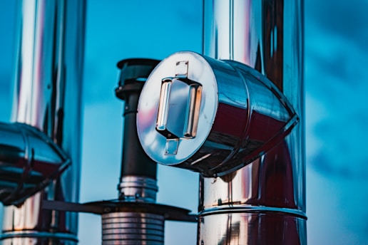 A close-up view of metallic industrial pipes reflecting light against a bright blue sky. The shiny surfaces highlight the cylindrical shapes and the detailed structure of the pipes. One prominent pipe with a circular cap is central to the image.