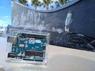 An Arduino UNO board is enclosed in a clear plastic case set against a background featuring a black stone wall with water fountains and an engraved image of a historical figure. Tall palm trees can be seen in the distance under a blue sky.