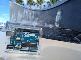 An Arduino UNO board is enclosed in a clear plastic case set against a background featuring a black stone wall with water fountains and an engraved image of a historical figure. Tall palm trees can be seen in the distance under a blue sky.