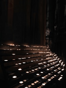 Rows of small lit candles are displayed on a multi-tiered stand inside a dimly lit space. The background is largely obscured, but hints of ornate architecture can be seen.
