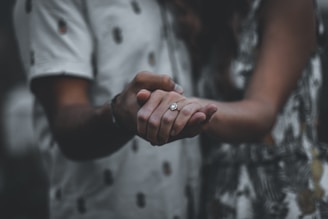 A couple holding hands, with a focus on a hand wearing a diamond ring. The background is softly blurred, creating an intimate and romantic atmosphere.