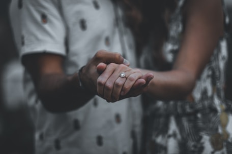 A couple holding hands, with a focus on a hand wearing a diamond ring. The background is softly blurred, creating an intimate and romantic atmosphere.