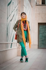 A person is standing in an urban setting wearing a colorful outfit. The attire includes a green coat, orange scarf, teal pants, and stylish sneakers. The background features a tiled path and a wall with a modern design.