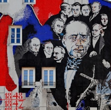 A large mural is painted on a building wall featuring historical figures dressed in 19th-century attire. The faces are depicted in a collage style with a mix of colors, primarily red, blue, black, and white. Two rectangular windows are integrated into the artwork. The mural seems to be divided into panels, with numbers like 1848 and 1847 painted at the bottom left corner.