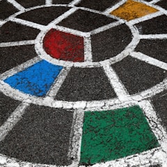 A painted section of pavement features a geometric design composed of white lines forming a segmented circular pattern. The segments are filled with different bright colors, including red, blue, green, and yellow, set against a dark asphalt background.