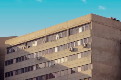 A multi-story concrete building with large windows is topped with air conditioning units on several floors. The façade shows signs of wear and shadow patterns from nearby structures. The sky above is clear with a soft blue tone.