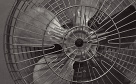 A close-up view of a metal fan with prominent blades and a protective wire cage. The image is in black and white, highlighting the metallic texture and structure of the fan. The focus is primarily on the center, where the blades converge behind the wire guard.