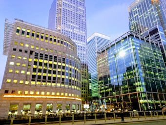 Tall modern skyscrapers with extensive use of glass and steel architecture dominate the scene. Bright lights from the office windows create a vibrant atmosphere, reflecting the bustling financial district setting. The sky is clear with a soft evening light.
