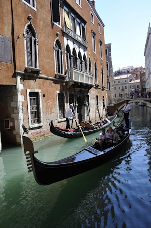 Gondolas glide along a narrow canal in Venice, Italy, with stone buildings featuring arched windows and balconies lining the waterway. Tourists are seated in the gondolas, enjoying the serene ride. A gondolier stands, expertly maneuvering his vessel with a long oar. In the background, a small bridge crosses the canal, connecting the historic structures.