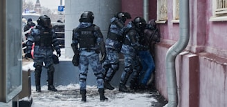 Several officers in riot gear are detaining an individual against a wall on a snowy street. The officers are dressed in dark uniform with helmets and protective armor. The setting appears to be urban with buildings and some people in the background.