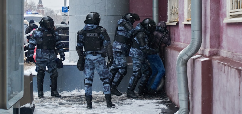 Several officers in riot gear are detaining an individual against a wall on a snowy street. The officers are dressed in dark uniform with helmets and protective armor. The setting appears to be urban with buildings and some people in the background.