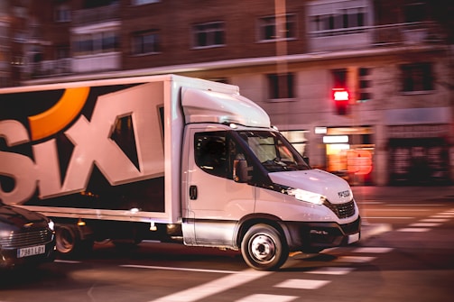 A white delivery truck with the word 'SIXT' displayed prominently on its side is driving through a city street. The environment suggests a motion blur effect, indicating speed and movement. Buildings and traffic lights are visible in the background.