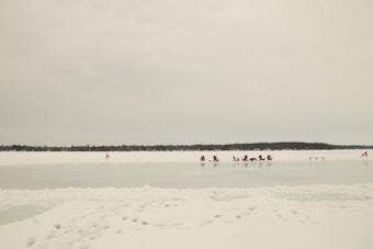 A series of red chairs are positioned on an expansive, frozen lake with snow covering the surface. Two small red flags are visible, one near the chairs and another at the edge of the scene. Trees are visible across the horizon under a cloudy sky.