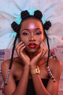 A woman with intricate braided hair styled with a large netted accessory. Her makeup is bold, with a glossy red lip and highlighted makeup. She is wearing a strapless top and a gold watch, posing with her hands gently touching her face.