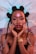 A woman with intricate braided hair styled with a large netted accessory. Her makeup is bold, with a glossy red lip and highlighted makeup. She is wearing a strapless top and a gold watch, posing with her hands gently touching her face.