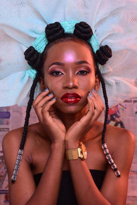 A woman with intricate braided hair styled with a large netted accessory. Her makeup is bold, with a glossy red lip and highlighted makeup. She is wearing a strapless top and a gold watch, posing with her hands gently touching her face.
