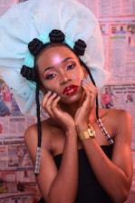 A woman with vibrant makeup and a large, intricate head wrap adorned with styled hair loops poses against a backdrop of newspapers. She holds her face gently with her hands, accentuating her elegant features and jewelry.