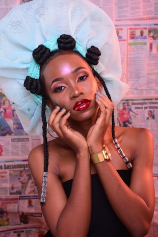 A woman with vibrant makeup and a large, intricate head wrap adorned with styled hair loops poses against a backdrop of newspapers. She holds her face gently with her hands, accentuating her elegant features and jewelry.