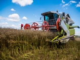 A large combine harvester is actively harvesting a field of tall crops under a partly cloudy blue sky. The machine is equipped with red rotating mechanisms and has branding on its side panel. The foreground features cut stalks, while the background shows standing crops.